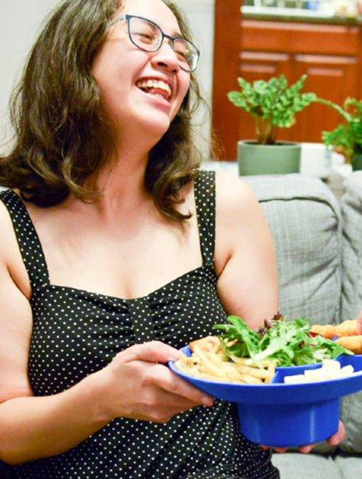 Bespectacled woman in a polka-dotted dress laughing while holding a bowl plate filled with chips