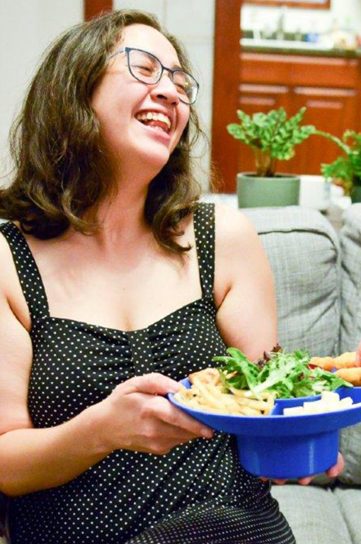 Bespectacled woman in a polka-dotted dress laughing while holding a bowl plate filled with chips