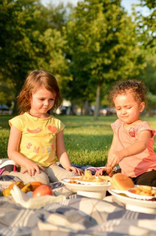 Two young girls sitting on the grass with a picnic blanket in front of them and two bowl plates filled with food on top of it